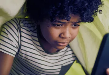 A child with curly hair, wearing a striped shirt, is intently looking at a smartphone under a blanket.