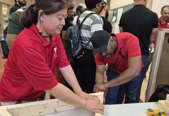 A woman holding wooden planks steady on a table as a man drills them together. There are several people in the background exploring skilled trades stations.