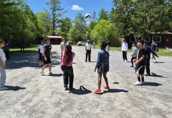 A group of newcomer youth playing with a volleyball on gravel. They are surrounded by a forested area.