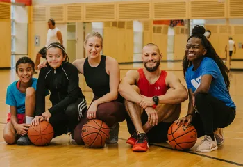 Group of five people sitting on a gym floor with basketballs, smiling at the camera. 