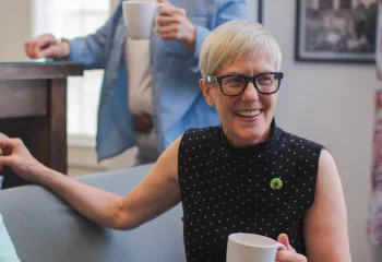 A senior woman sitting on a couch, holding a coffee mug and smiling. There are other folks around her.