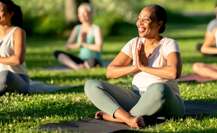 A woman smiles while sitting cross-legged on a yoga mat outdoors, hands in prayer position.
