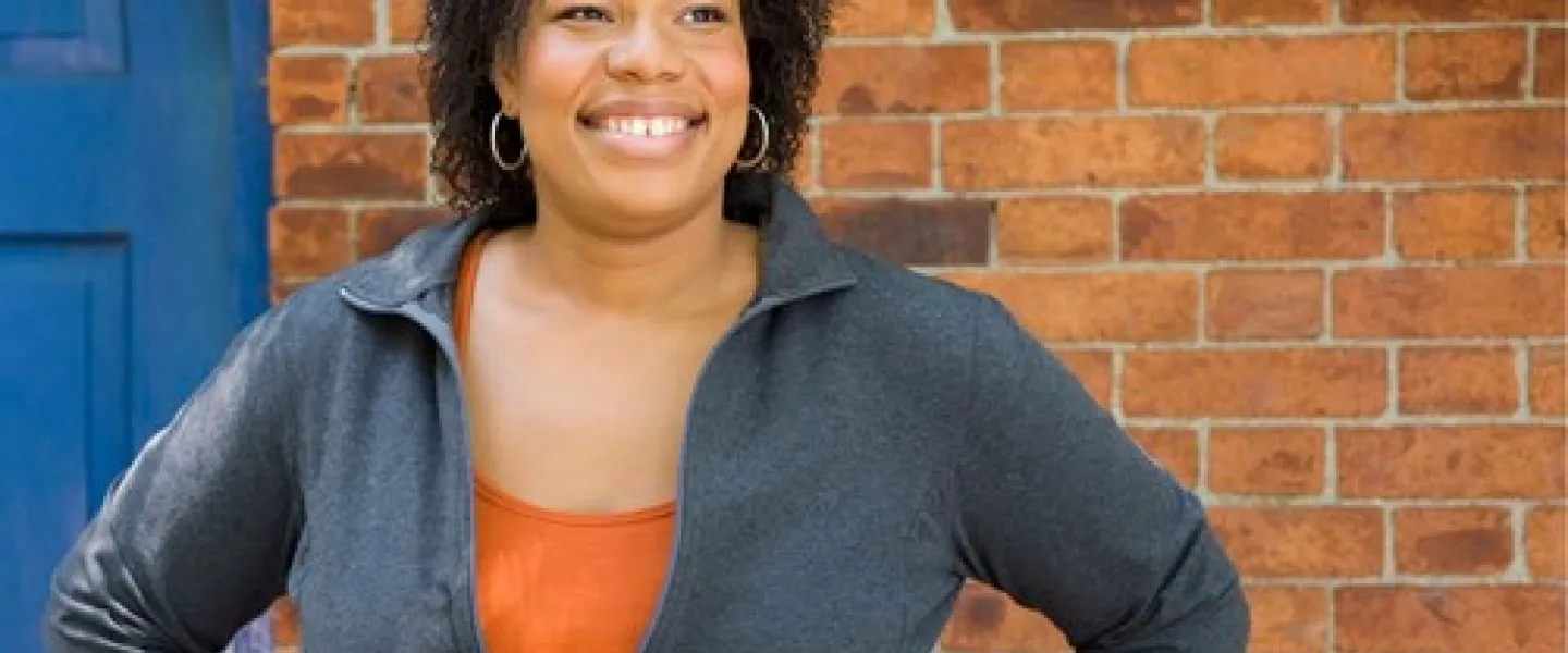 A black woman smiling with her hands on her hips. She is standing in front of a brick wall and blue door.