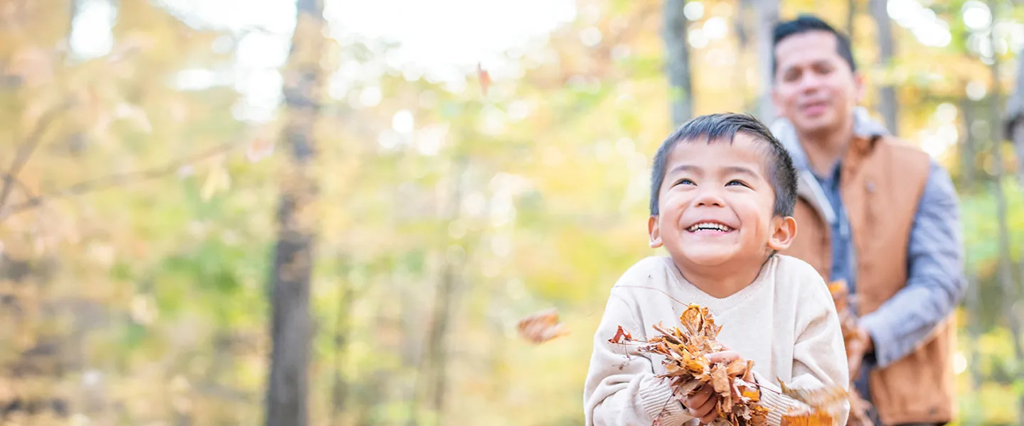 A man and a child smile while holding colorful leaves in a sunny outdoor setting.