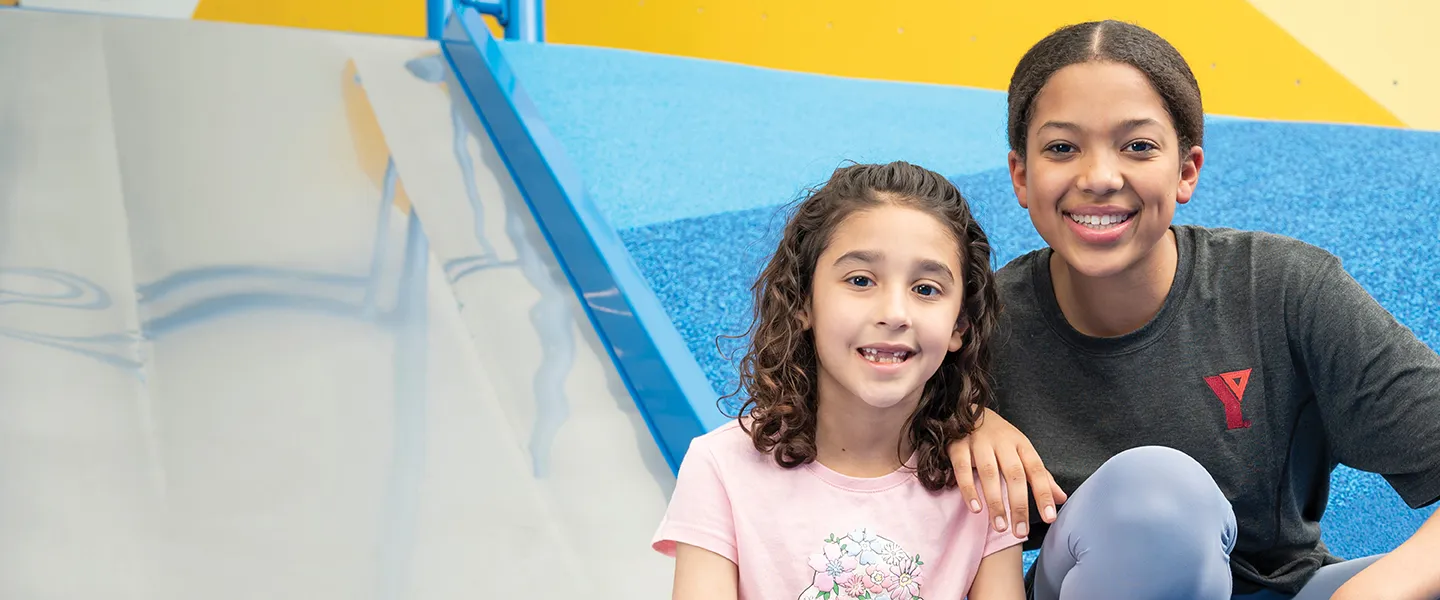 Two young girls sitting on a slide in a gym, smiling and enjoying their playtime together.