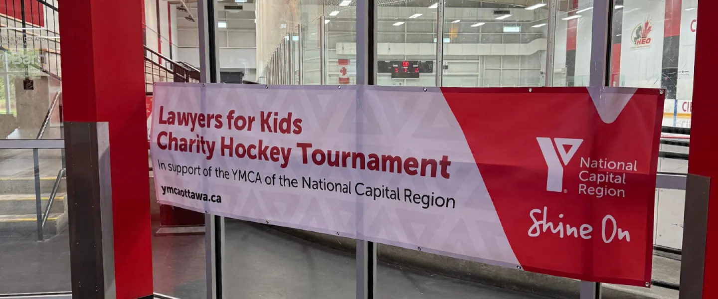 A red and white banner hanging on a glass wall in front of an ice rink. The banner reads: Lawyers for Kids Charity Hockey Tournament in support of the YMCA of the National Capital Region.