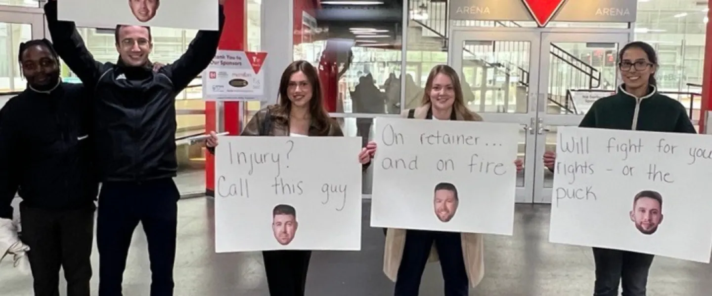 Five spectators holding up four signs with the faces of hockey team players. The signs read: "Billing hours, killing powerplays", "Injury? Call this guy", "On retainer... and on fire", "Will fight for your rights - or the puck."