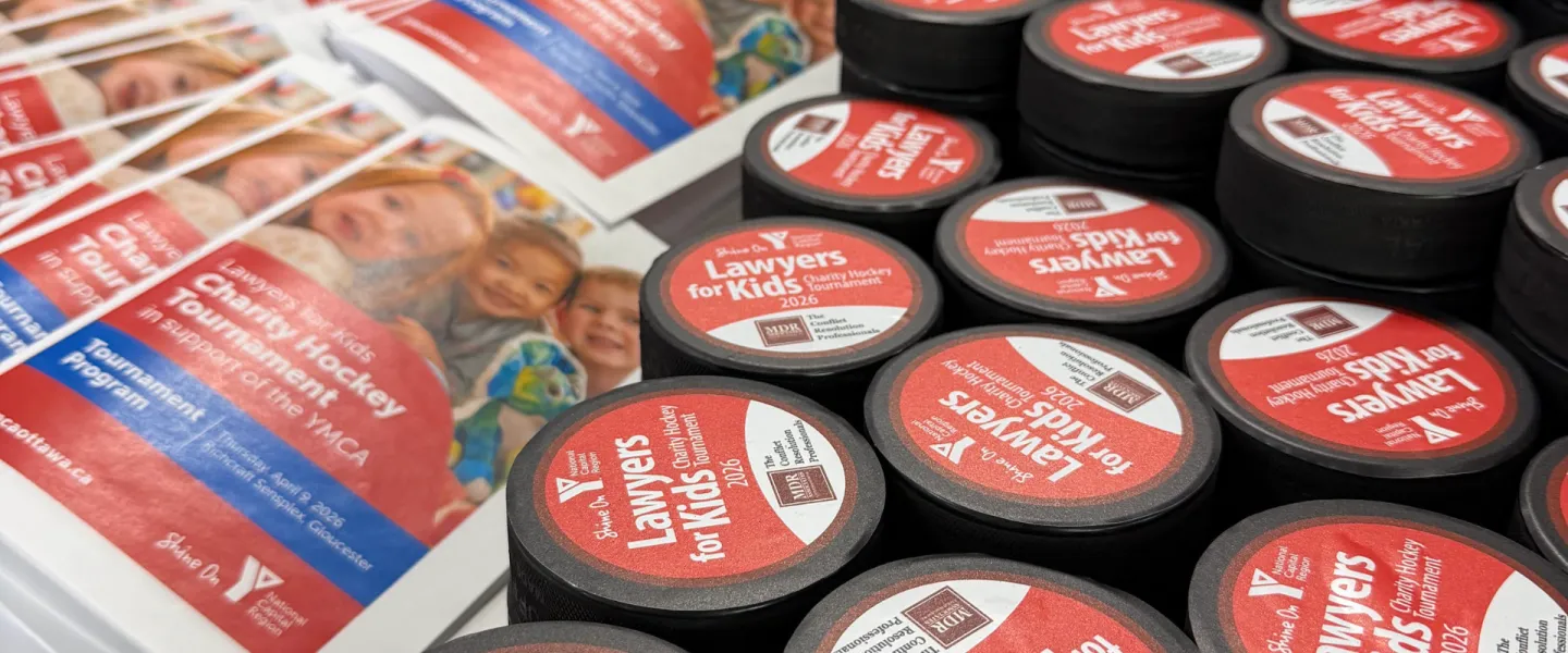 A close up of Lawyers for Kids Charity Hockey Tournament pamphlets, next to several hockey pucks that have Lawyers for Kids printed on them