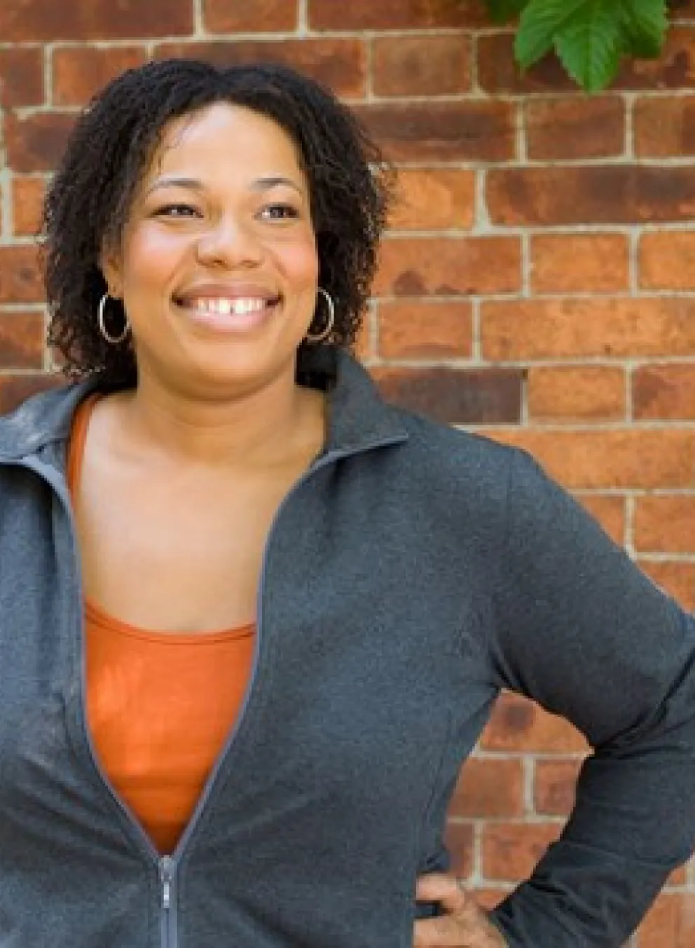 A black woman smiling with her hands on her hips. She is standing in front of a brick wall and blue door.