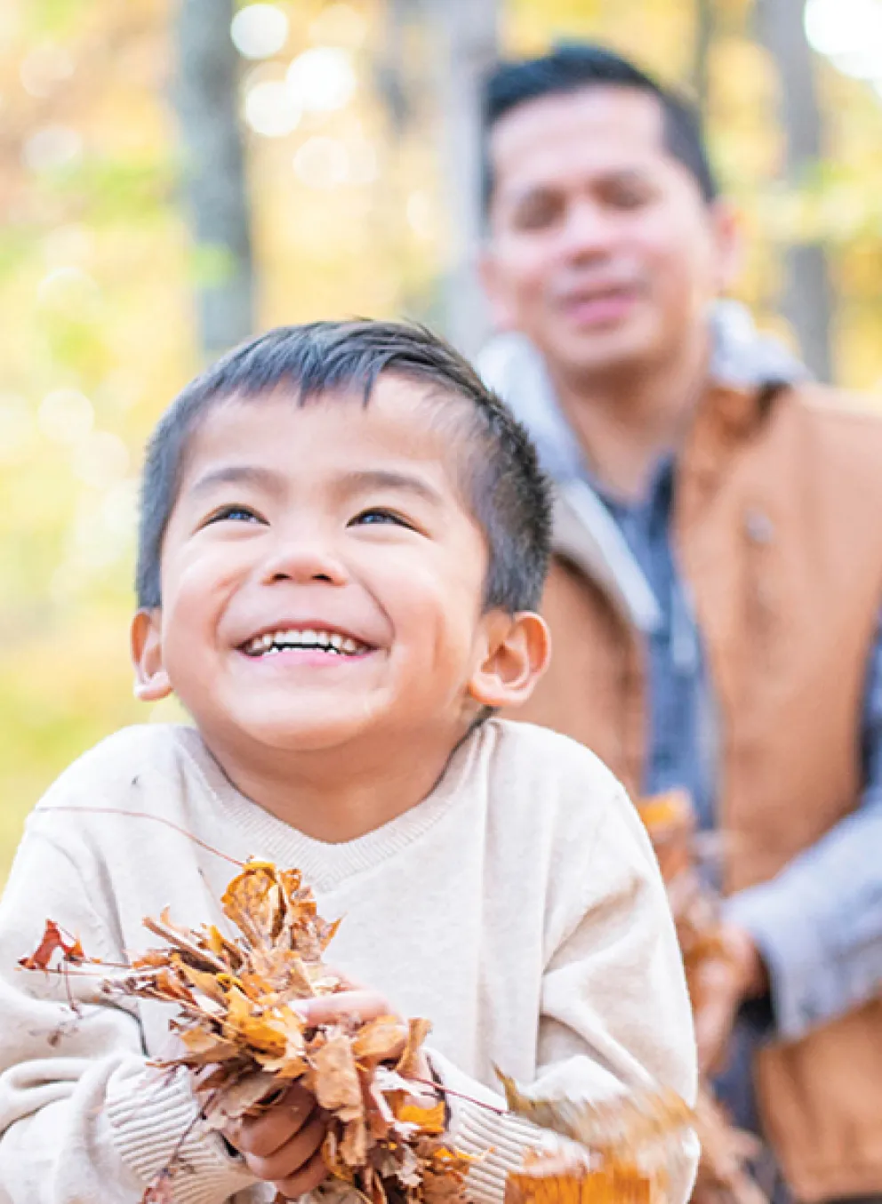 A man and a child smile while holding colorful leaves in a sunny outdoor setting.