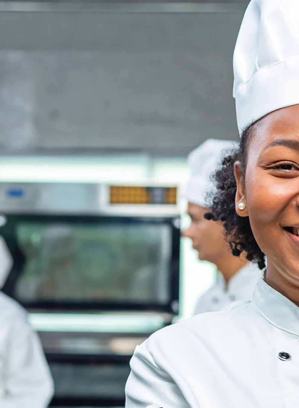 Three chefs in white uniforms and hats stand in a commercial kitchen, with one chef smiling confidently at the front of the image