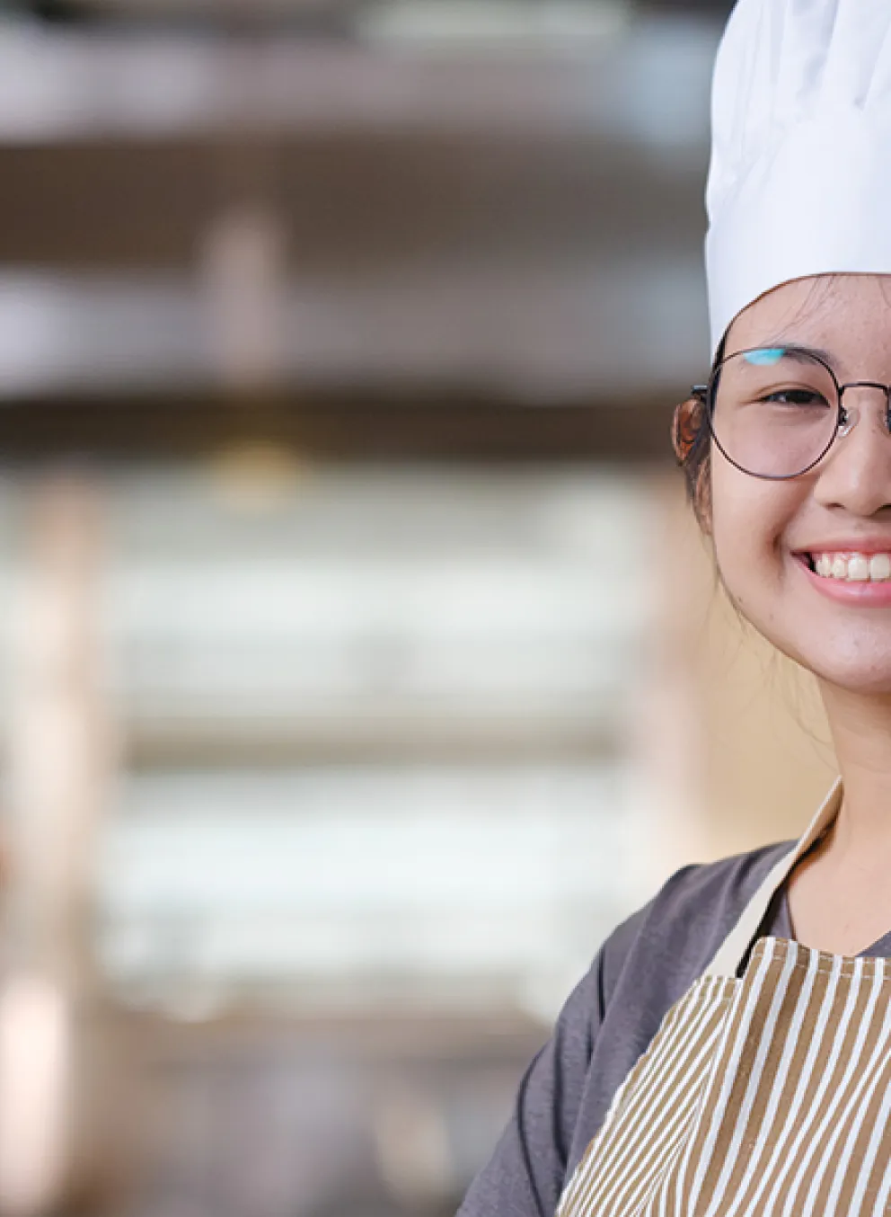 A young chef in a striped apron and white chef hat smiles at the camera in a commercial kitchen, with another chef working in the background.