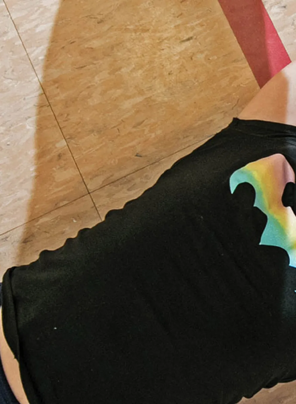 Young girl lies on a gym floor with arms outstretched, smiling and wearing a black shirt with a rainbow bat symbol.