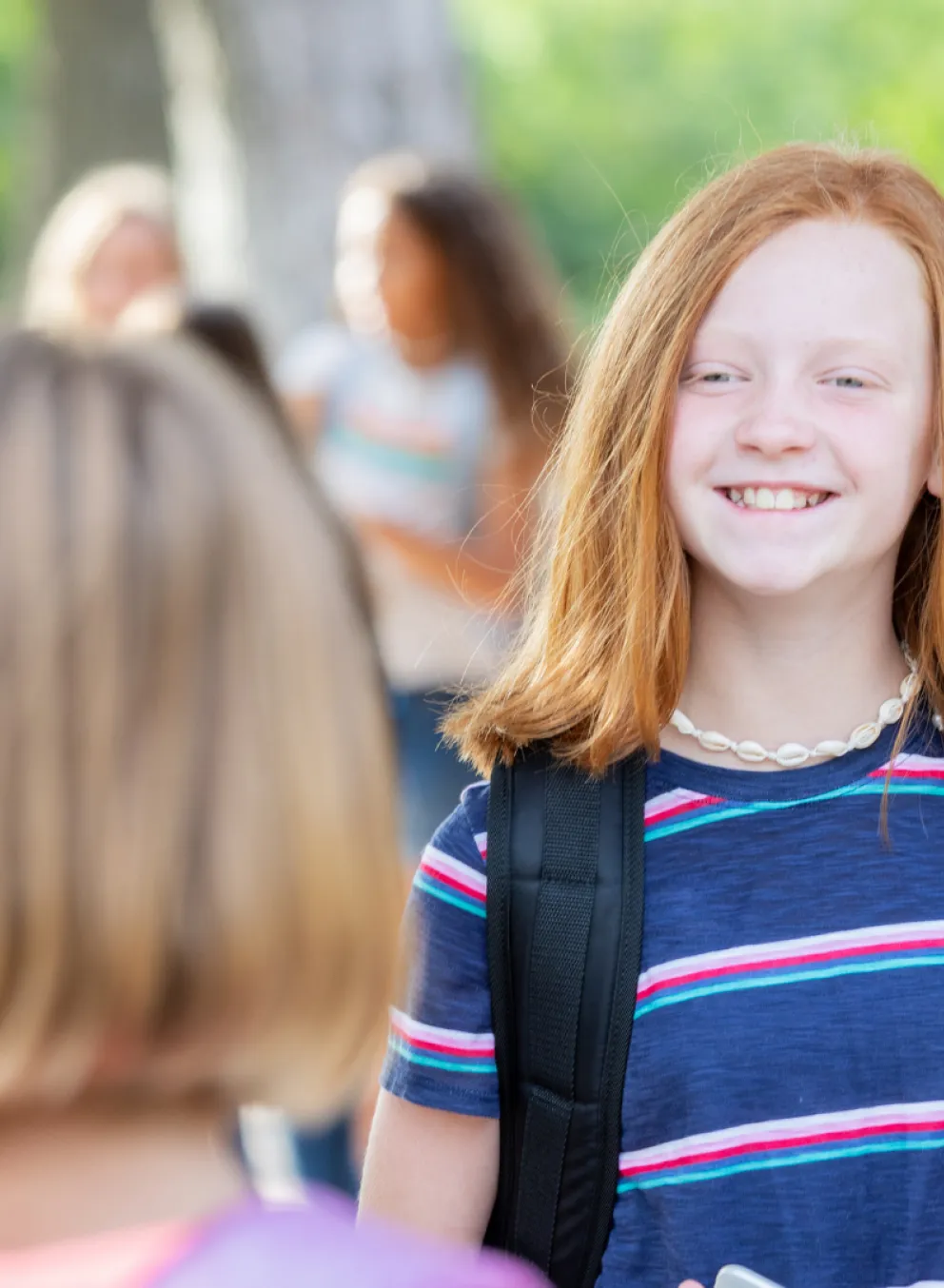 Two girls smiling at each other, wearing backpacks in a bright, outdoor setting