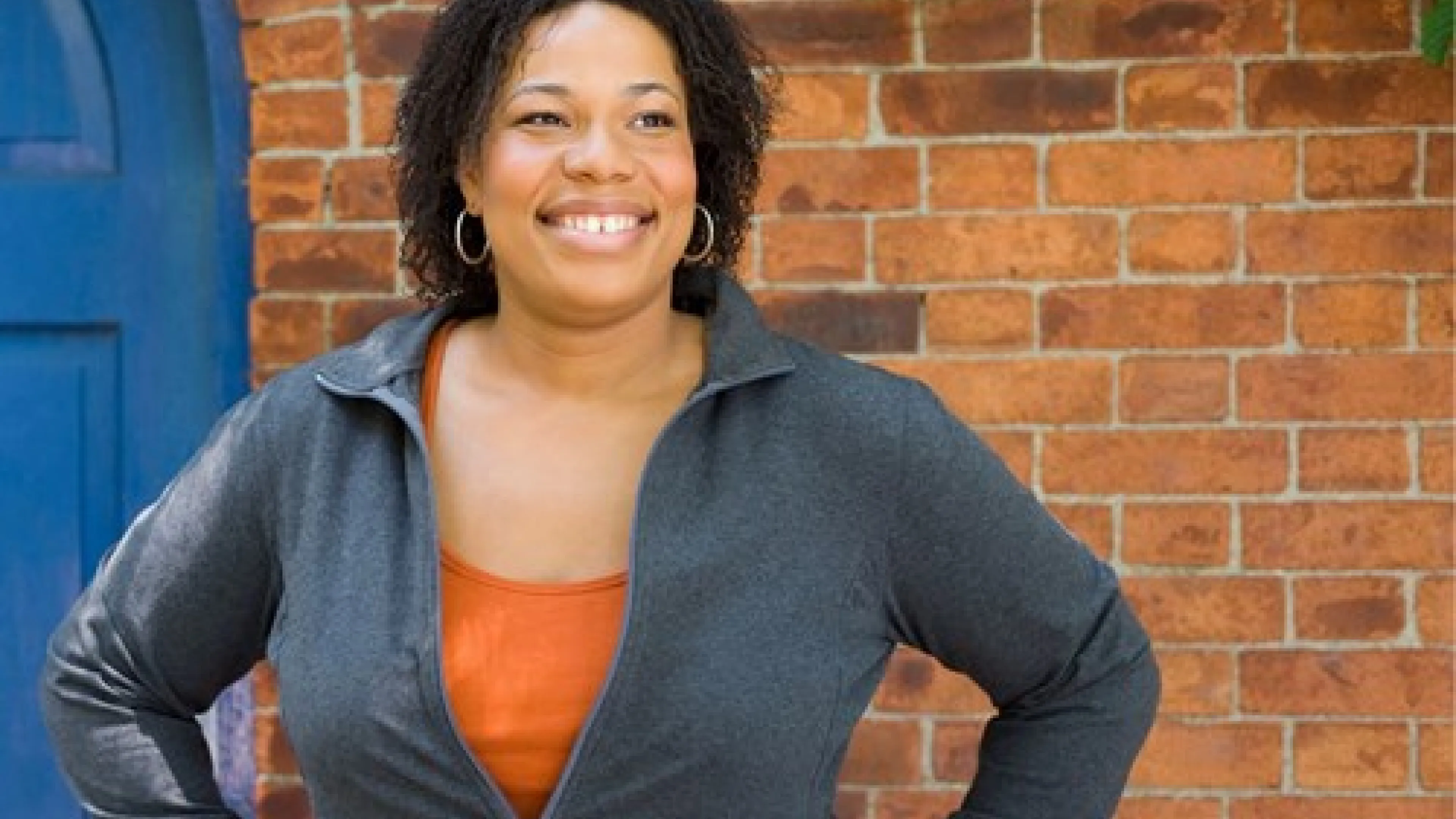 A black woman smiling with her hands on her hips. She is standing in front of a brick wall and blue door.