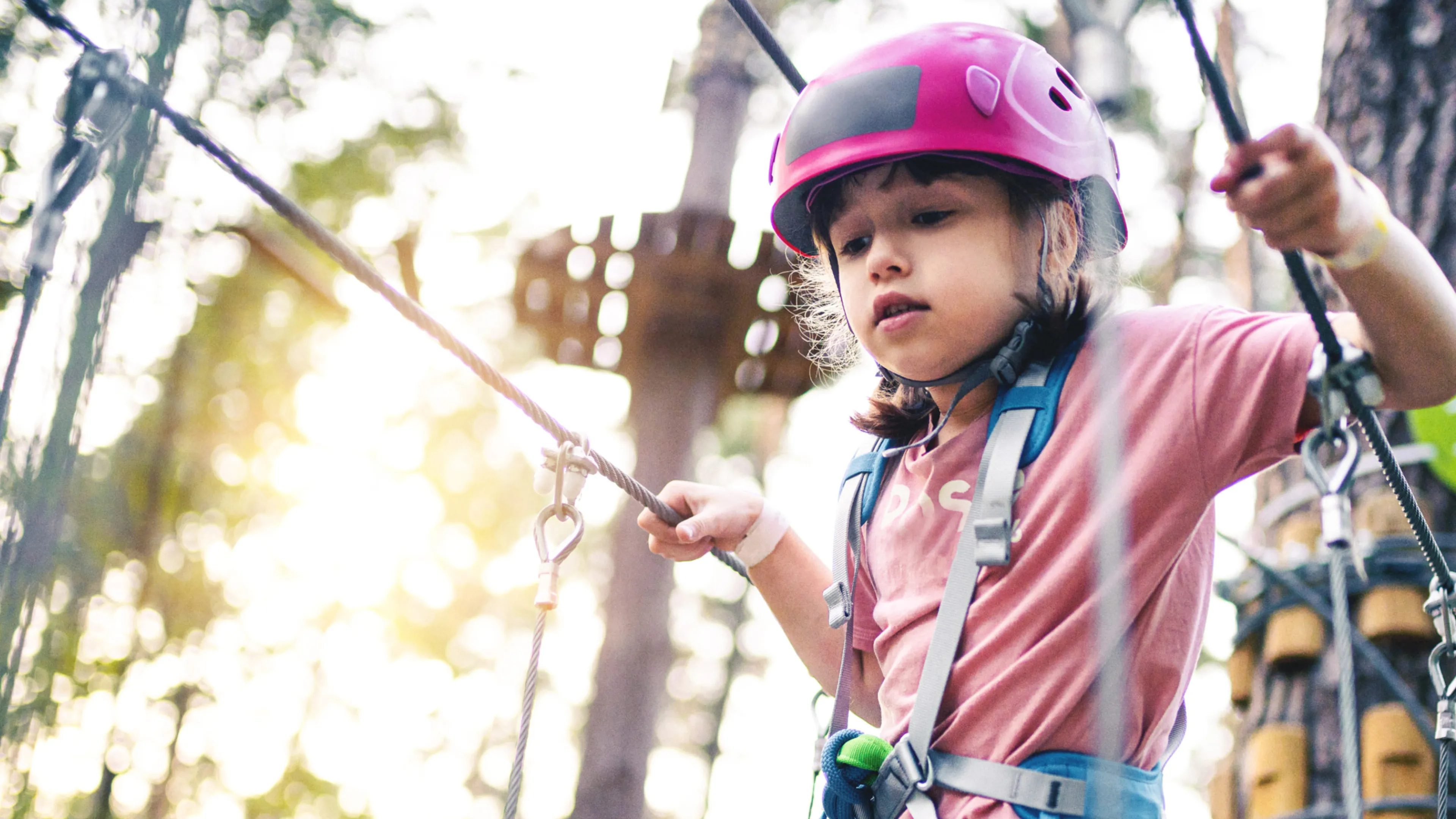 A girl wearing a helmet and safety harness navigates a high ropes course.