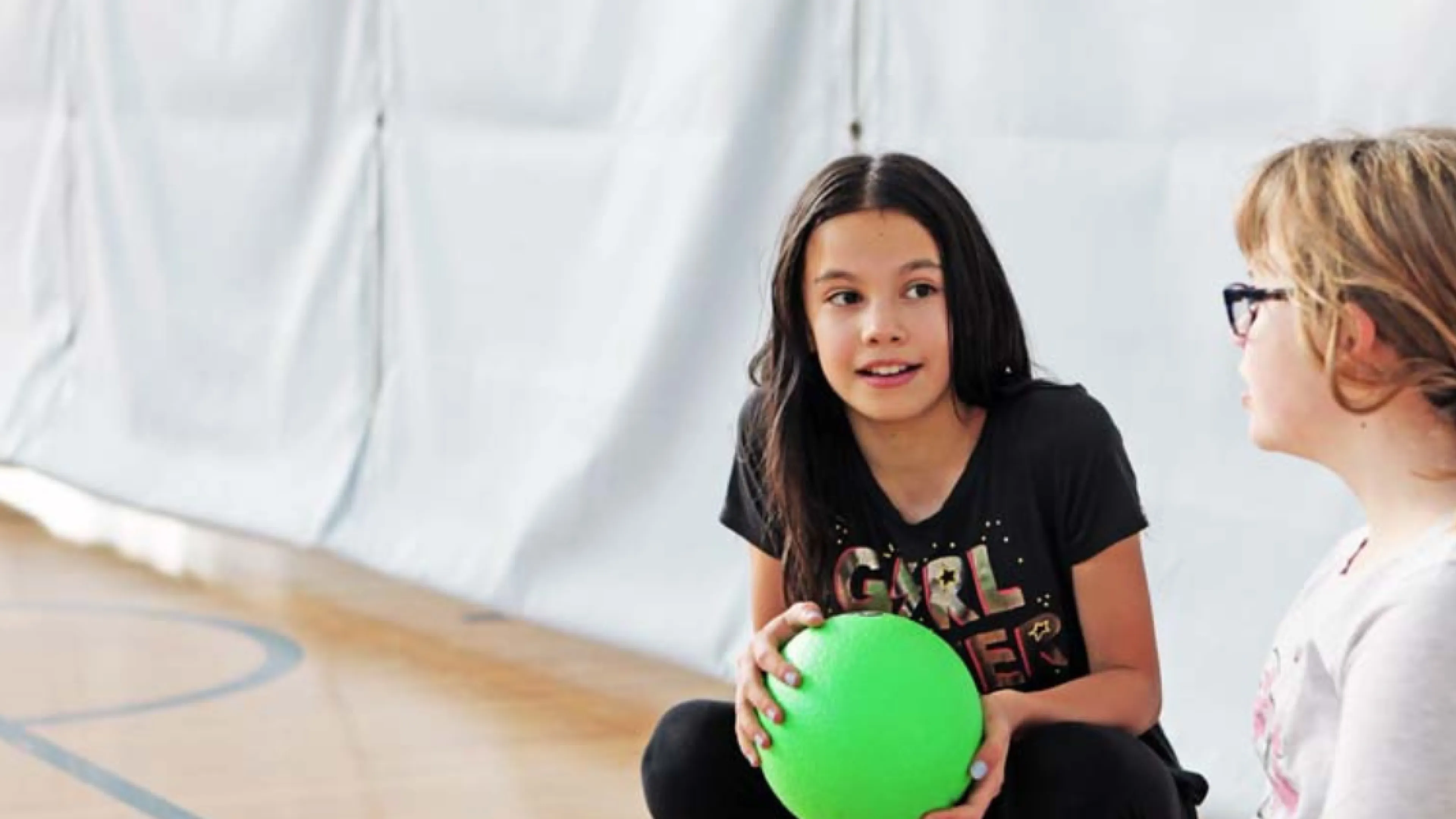Two young girls sit in a gymnasium with a  ball.