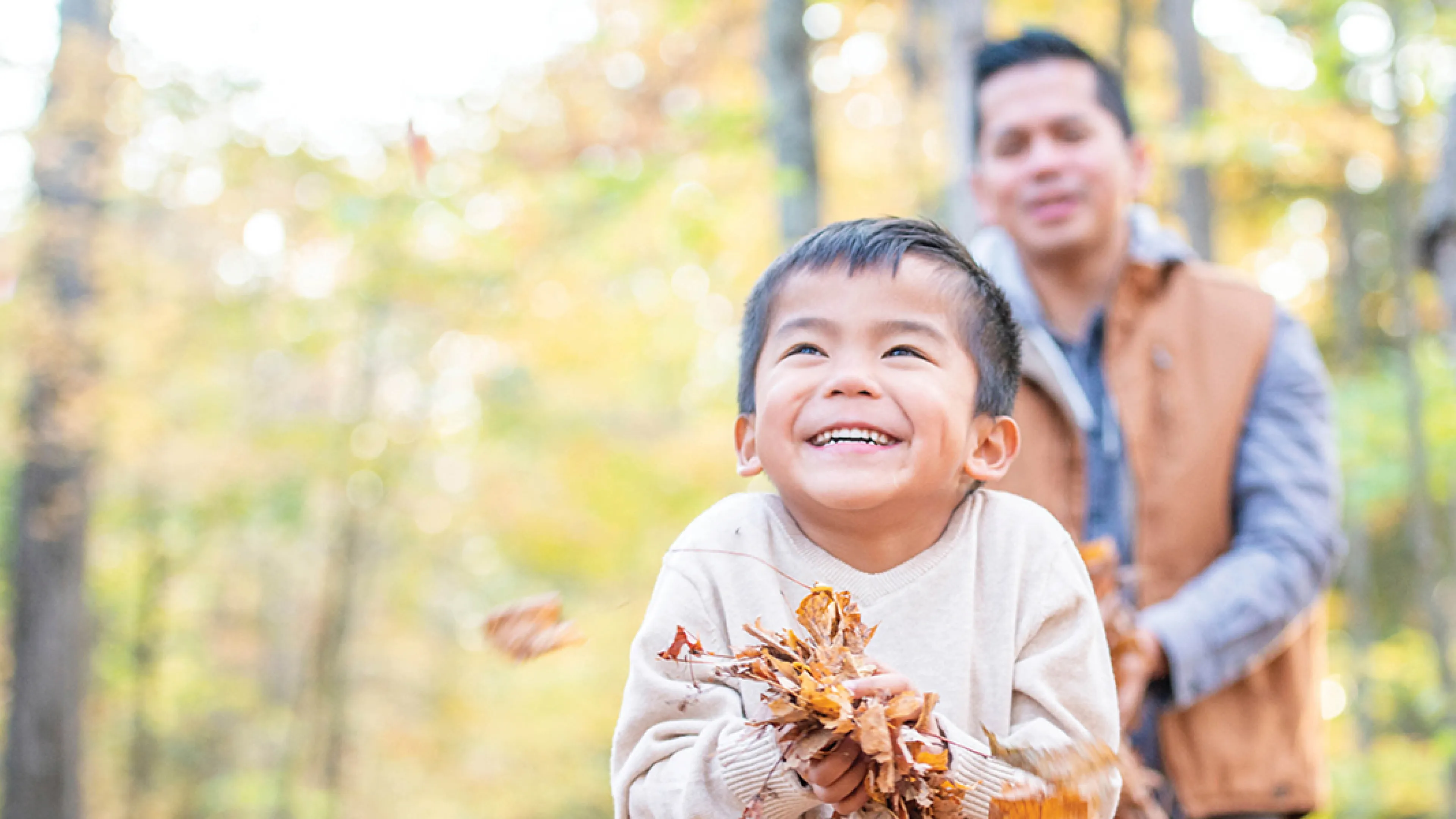 A man and a child smile while holding colorful leaves in a sunny outdoor setting.