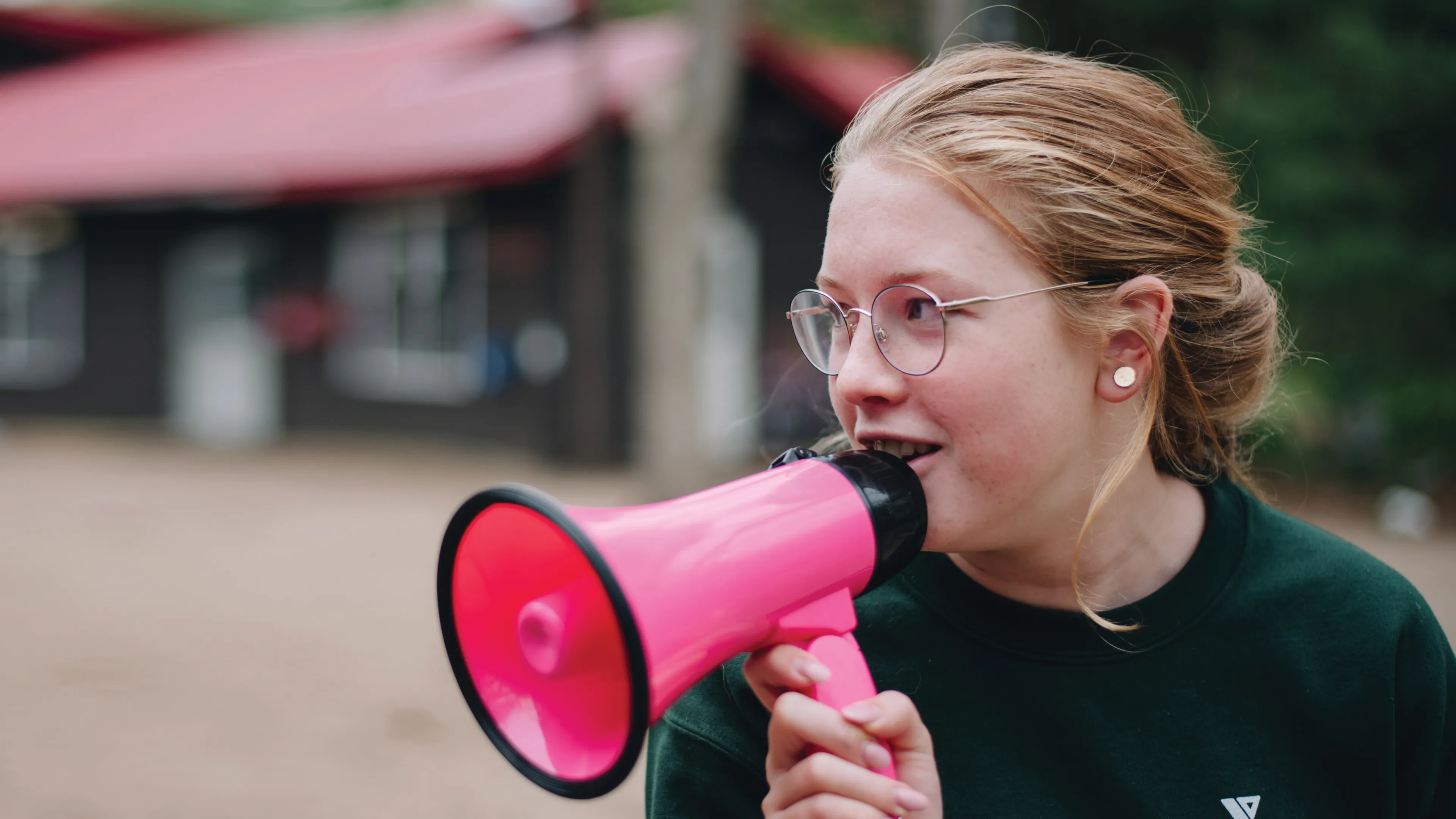 A girl with glasses enthusiastically holds a pink megaphone, ready to speak or cheer.