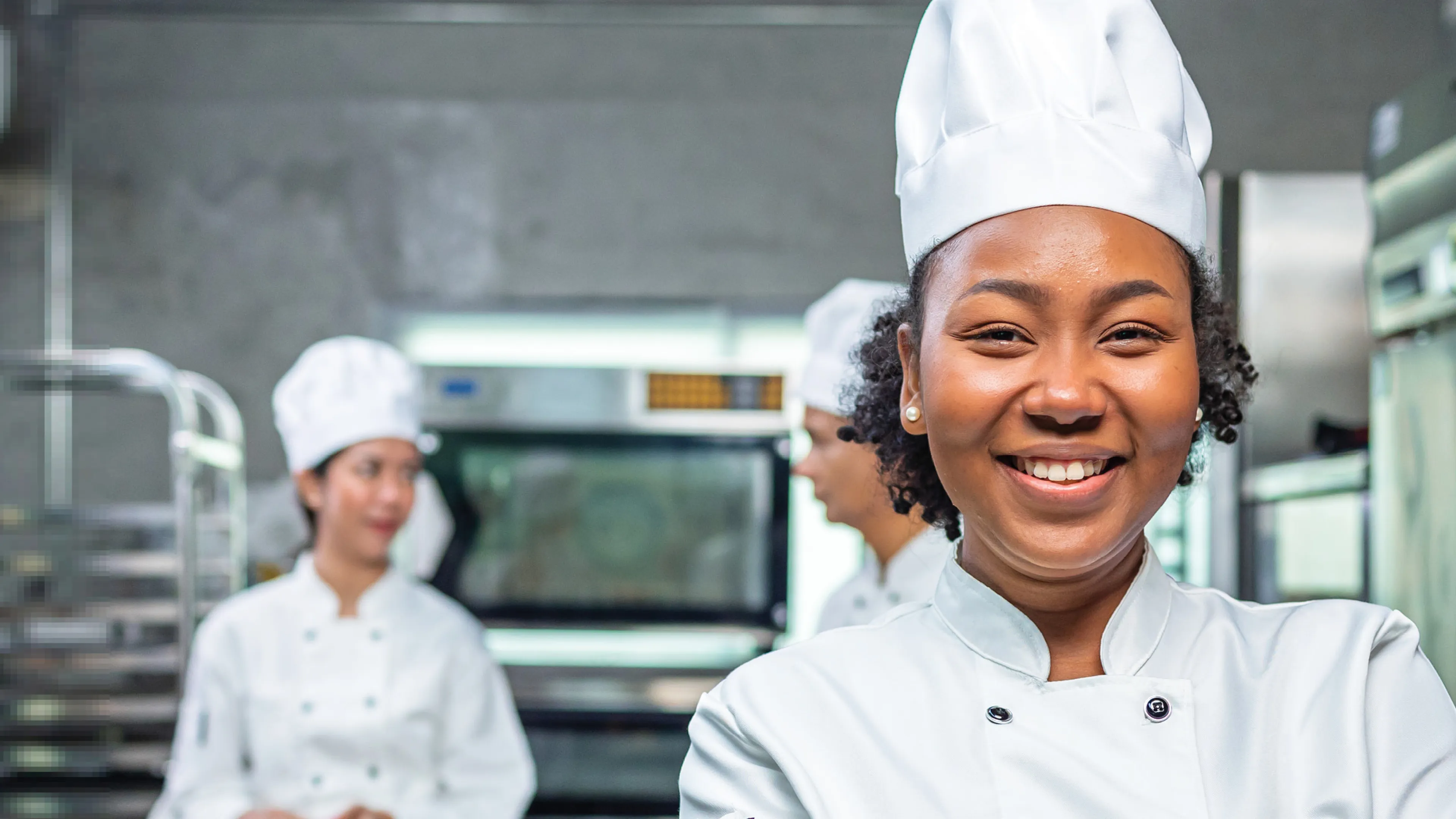 Three chefs in white uniforms and hats stand in a commercial kitchen, with one chef smiling confidently at the front of the image