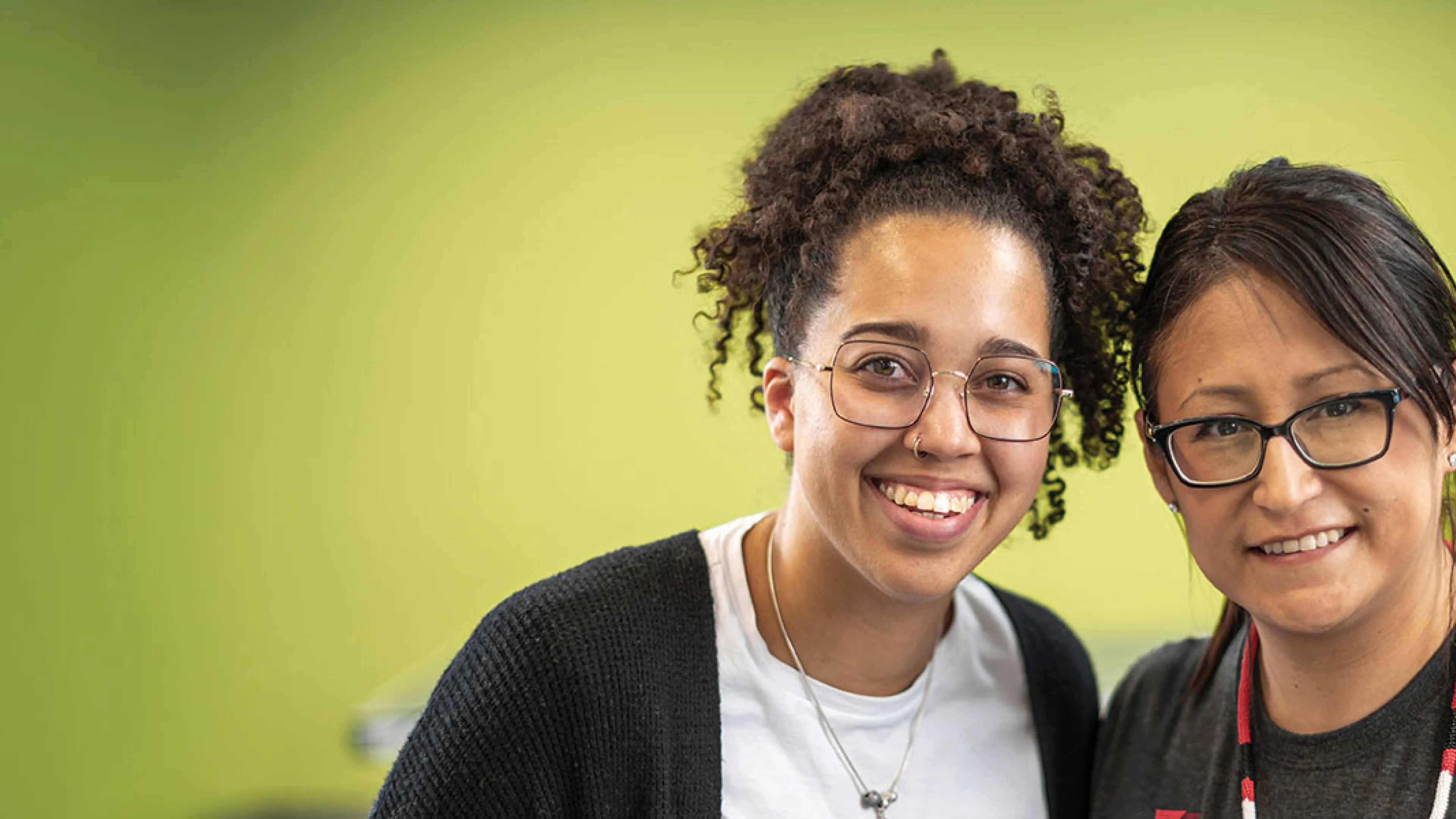Two smiling women wearing glasses, posing together against a green background.