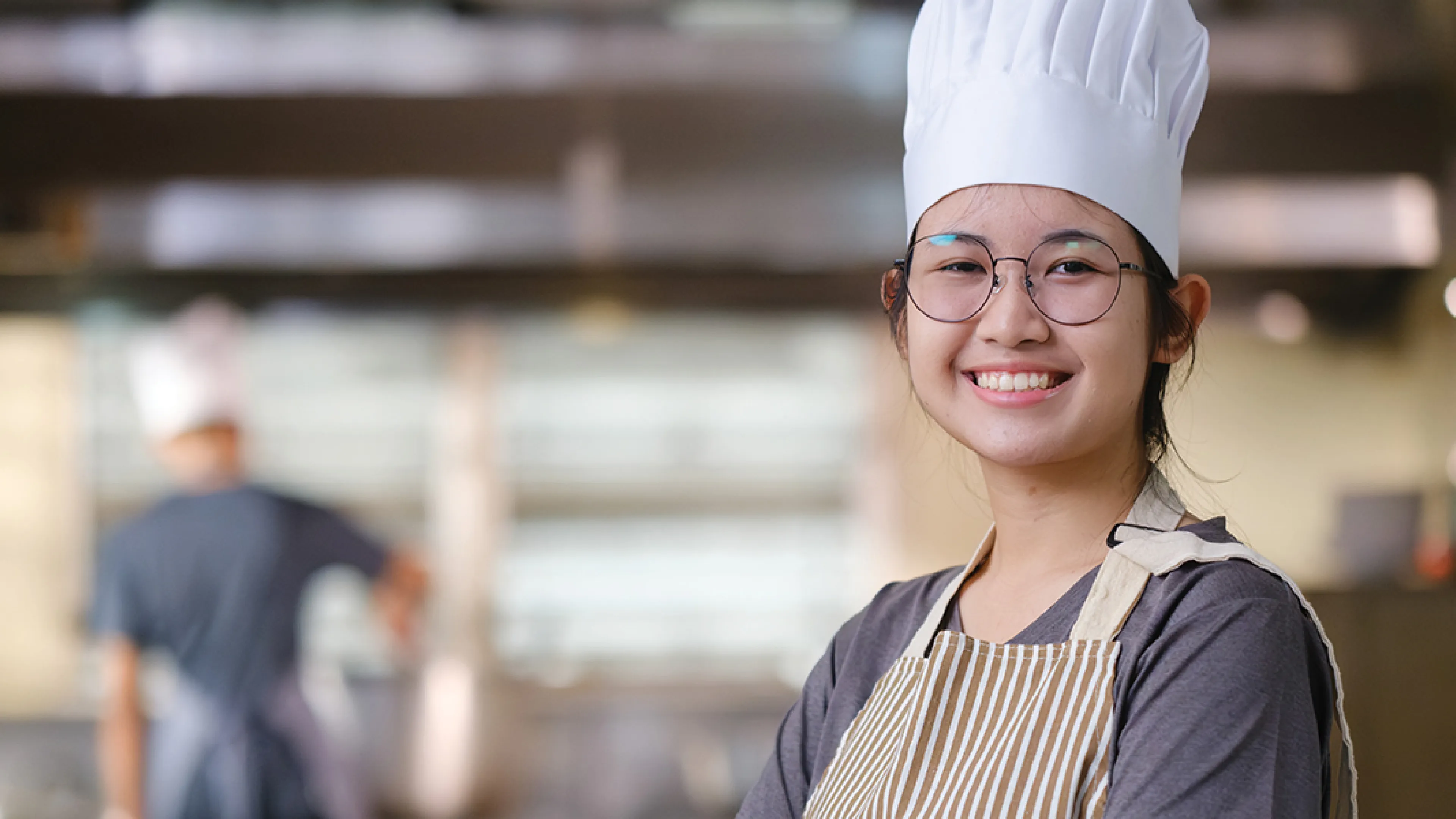 A young chef in a striped apron and white chef hat smiles at the camera in a commercial kitchen, with another chef working in the background.