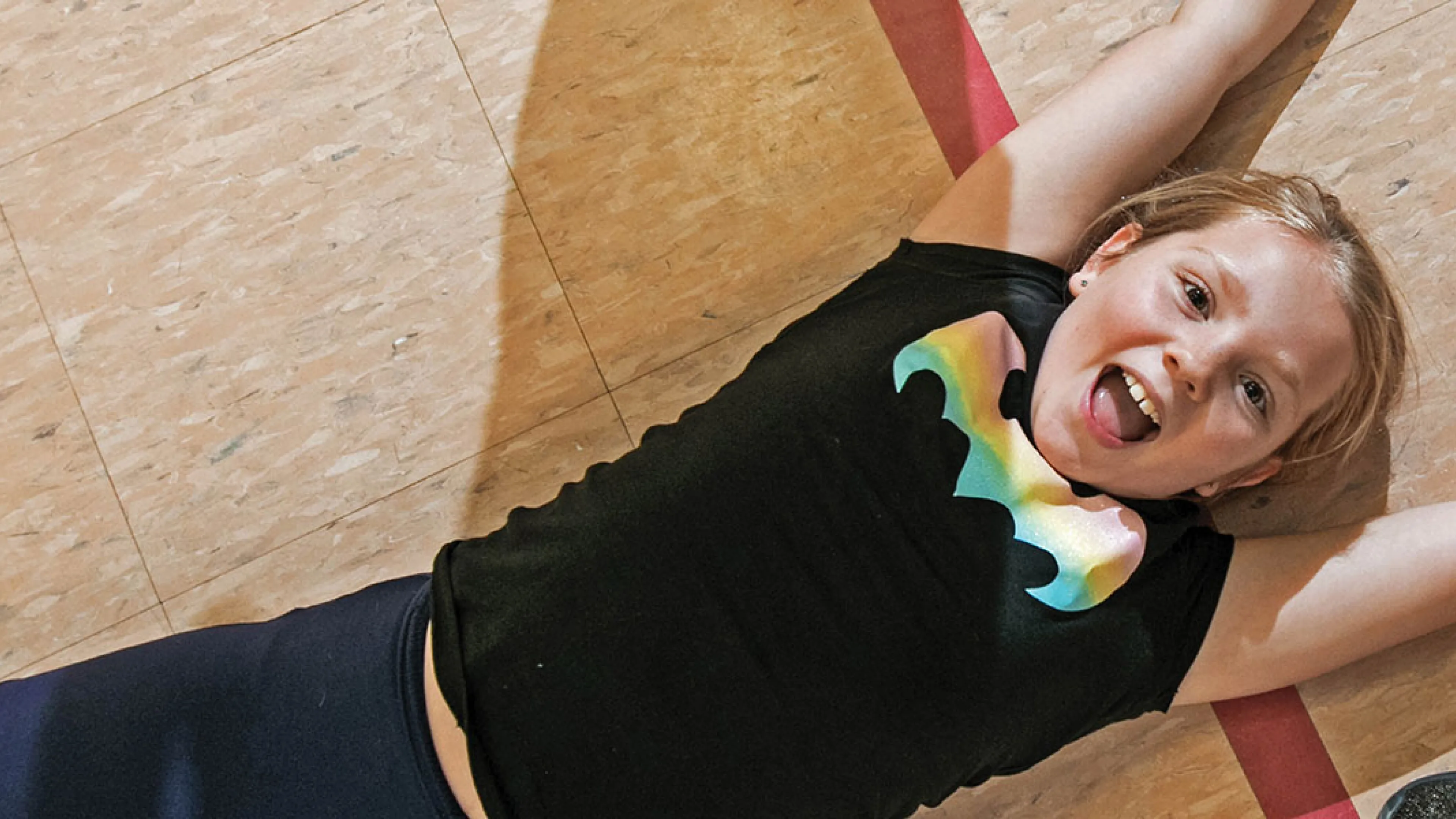 Young girl lies on a gym floor with arms outstretched, smiling and wearing a black shirt with a rainbow bat symbol.