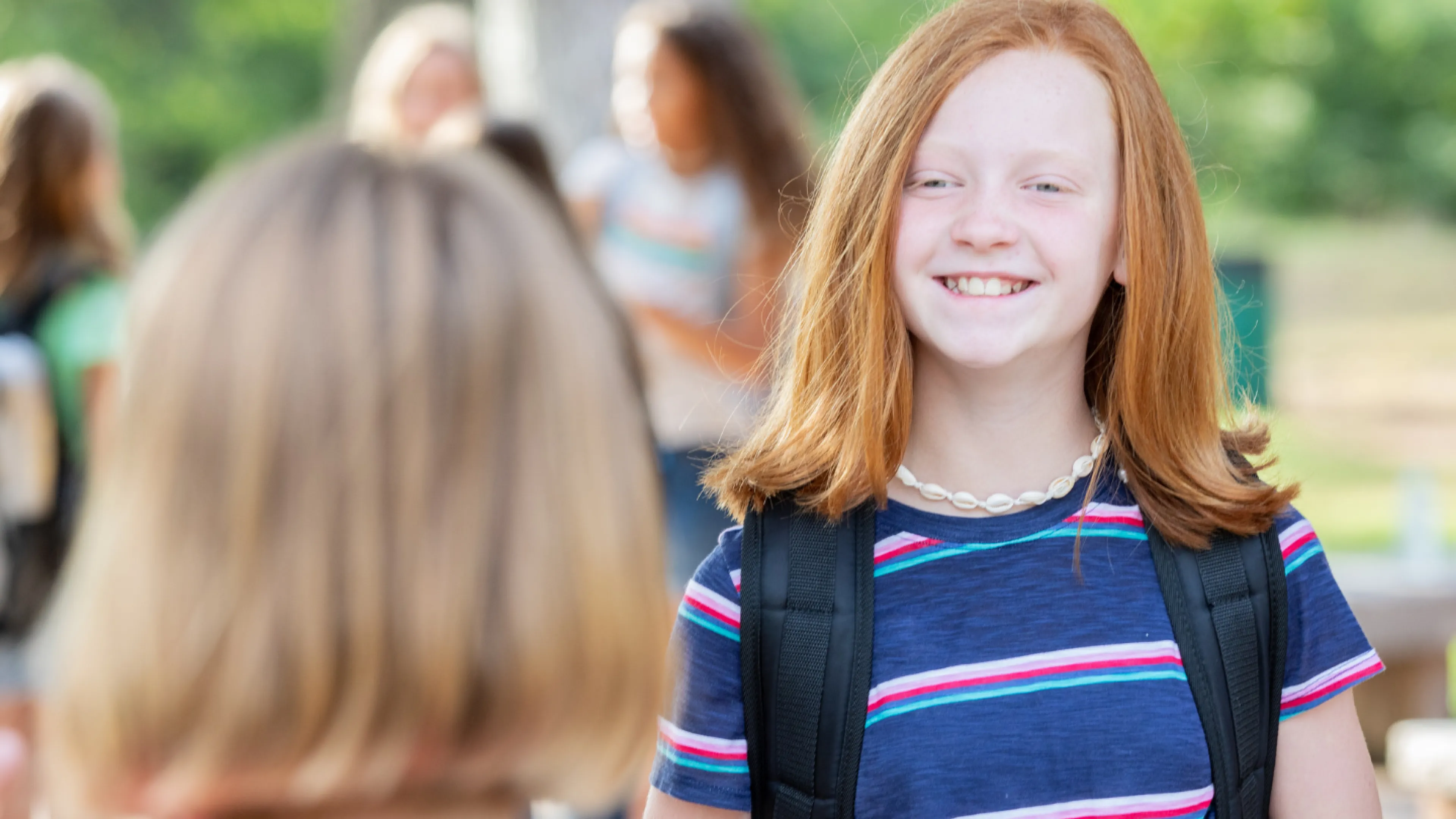 Two girls smiling at each other, wearing backpacks in a bright, outdoor setting
