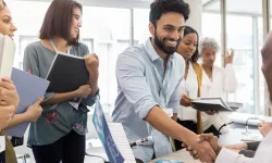 A diverse group of people in a job fair setting. A man in the foreground is smiling, shaking hands, and surrounded by others holding folders.