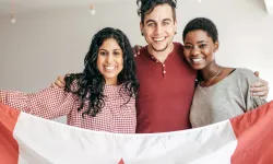 A man and two women, all of different cultural backgrounds, smiling and holding up a Canadian flag together