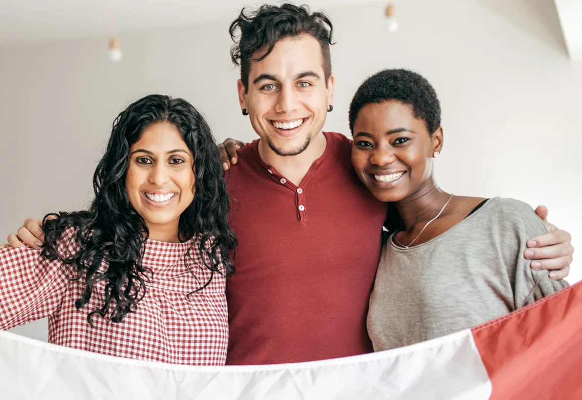 Three people stand close together indoors, smiling at the camera, with their arms around each other and holding a red and white flag.
