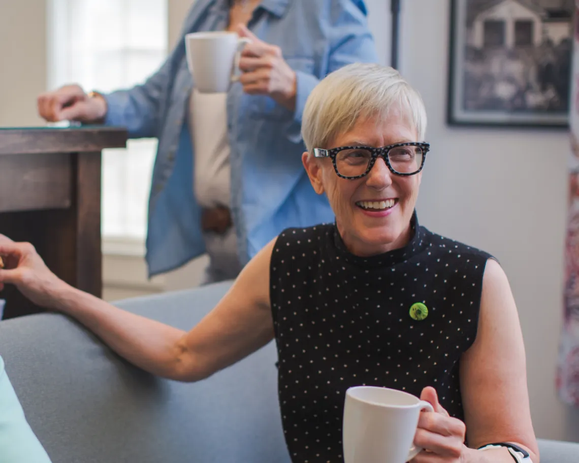 A senior woman sitting on a couch, holding a coffee mug and smiling. There are other folks around her.