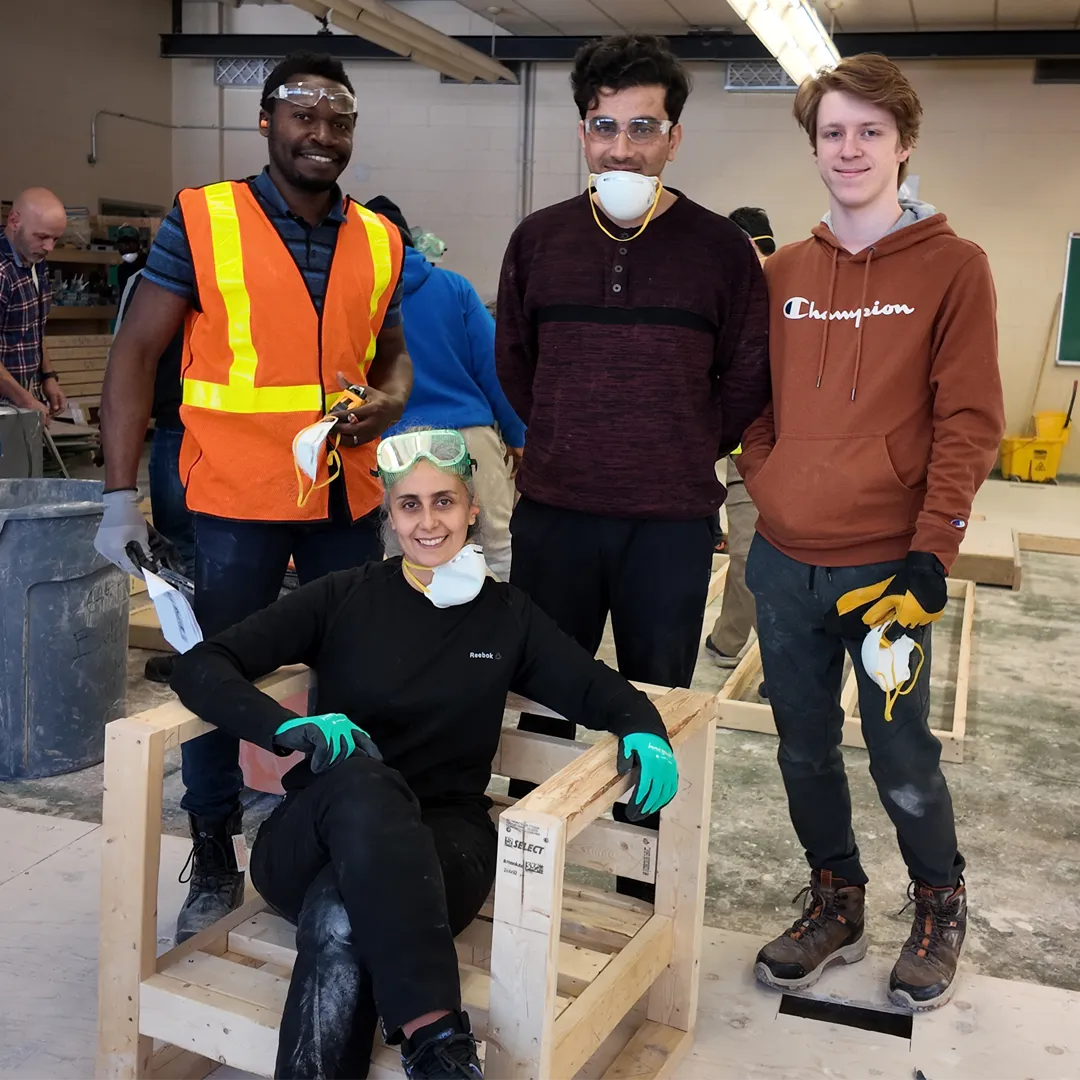 Four smiling workshop participants pose among unfinished wooden frames; one sits in a frame wearing safety goggles and gloves, while others wear protective gear.