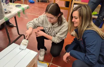 A teen camper painting a wooden bench while her aunt sits next to her