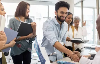 A diverse group of people in a job fair setting. A man in the foreground is smiling, shaking hands, and surrounded by others holding folders.