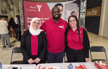 Three YMCA staff members standing and smiling together behind a booth promoting YMCA Employment and Newcomer Services at Merivale Mall.