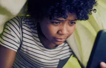 A child with curly hair, wearing a striped shirt, is intently looking at a smartphone under a blanket.