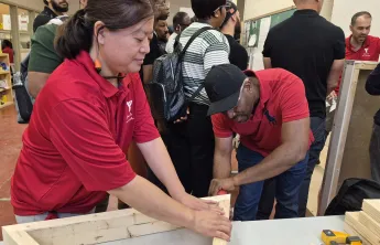 A woman holding wooden planks steady on a table as a man drills them together. There are several people in the background exploring skilled trades stations.