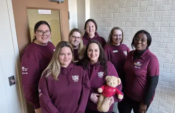 Group photo of seven YMCA Child Care team members wearing burgundy hoodies. One team member is holding a teddy bear wearing a Y Shine On shirt.