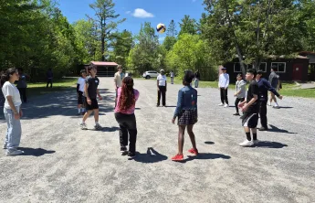 A group of newcomer youth playing with a volleyball on gravel. They are surrounded by a forested area.