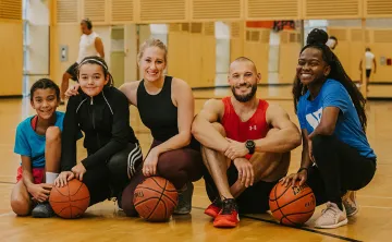 Group of five people sitting on a gym floor with basketballs, smiling at the camera. 