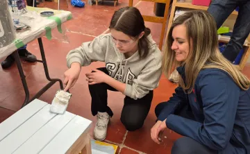 A teen camper painting a wooden bench while her aunt sits next to her