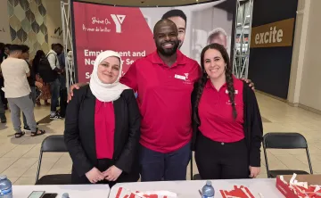 Three YMCA staff members standing and smiling together behind a booth promoting YMCA Employment and Newcomer Services at Merivale Mall.
