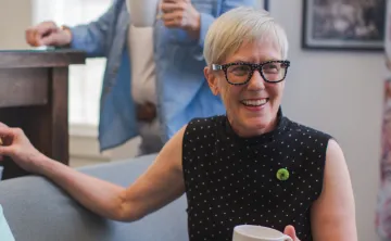 A senior woman sitting on a couch, holding a coffee mug and smiling. There are other folks around her.